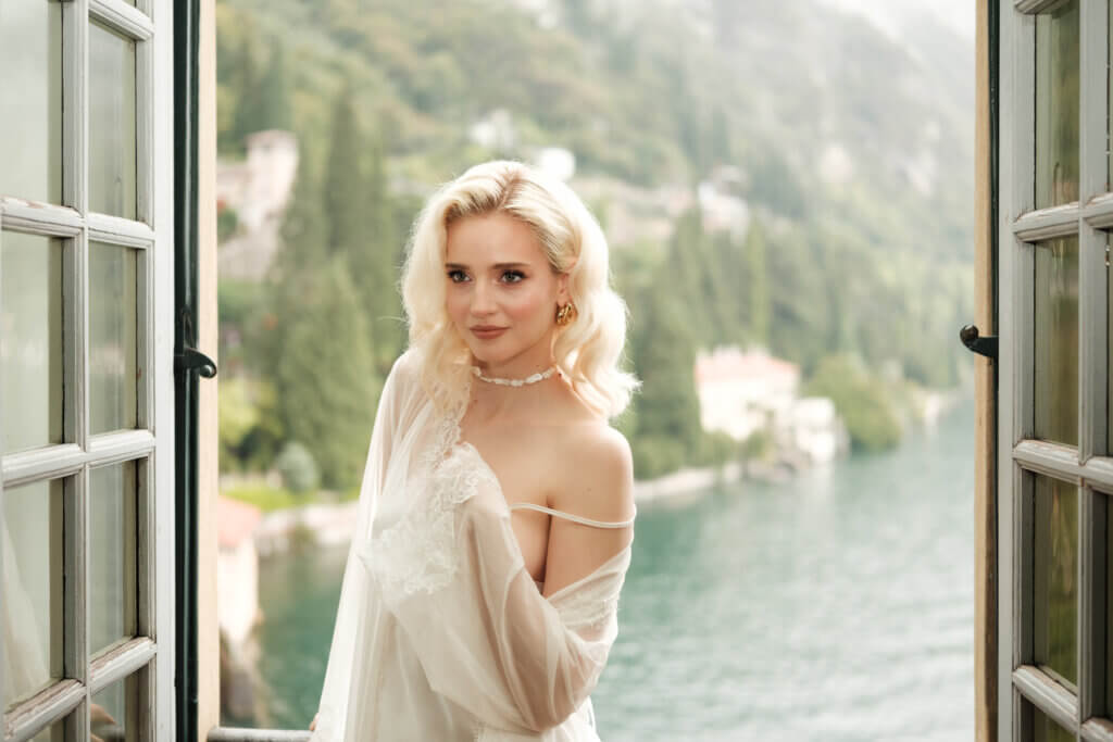 Blonde Braut im zarten Kleid am geöffneten Fenster mit Blick auf einen See