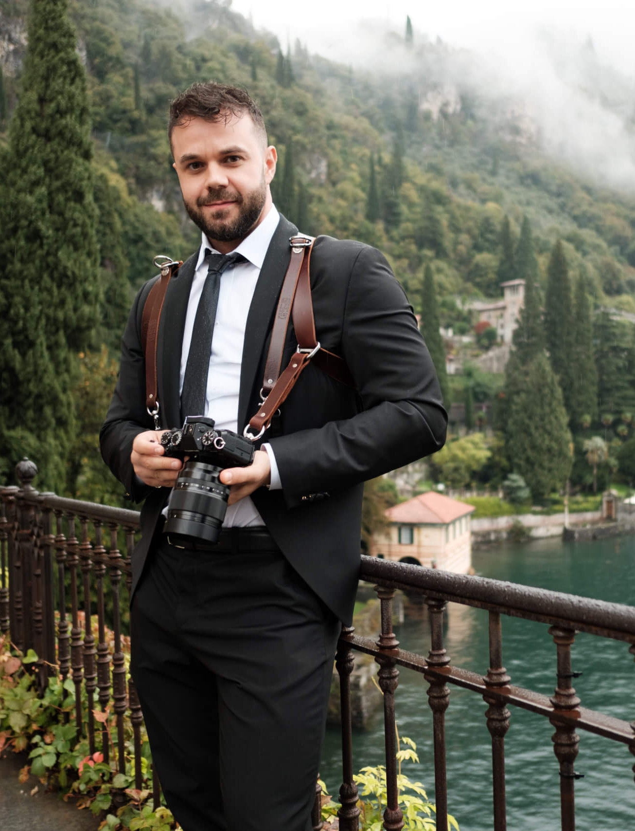 Agent Qufaj, Hochzeitsfotograf, im Anzug mit Kameragurt und Kamera an einer Seepromenade